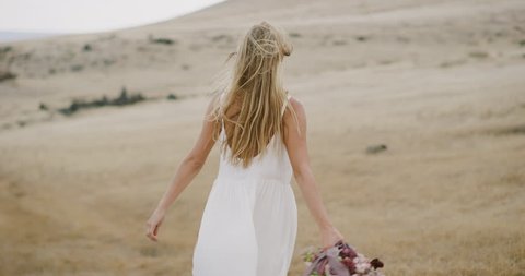 Beautiful young woman in a white dress smiling and twirling in a golden field holding a flower bouquet with her hair blowing in the wind in slow motion, bohemian bride lifestyle