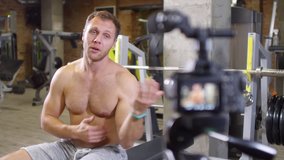 Shirtless muscular coach sitting on bench in gym, doing one arm dumbbell curls and then explaining how muscles work during exercise while recording video lesson with professional camera
