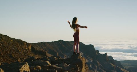 Young athletic woman standing on the edge of a cliff with her arms in the air watching an epic sunrise, amazing outdoors fitness lifestyle