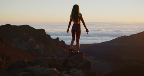 Young athletic woman raising her arms into the air in success pose while she looks out at epic sunrise from a mountaintop, amazing outdoors fitness in slow motion