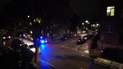 Police lights flash in the night after a disturbance in a residential road of West London. A taxi cab drives by.