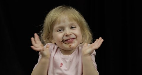 Beautiful, happy child with dirty face from melted chocolate and whipped cream on black background. Teen blonde girl in pink shirt making faces. Happy childhood. Joyful cute smile after eating sweets
