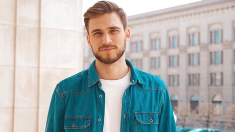 Portrait of handsome smiling stylish hipster lumbersexual model. Man dressed in jeans jacket clothes. Fashion male posing on the street background. Touching his hair. Slow motion