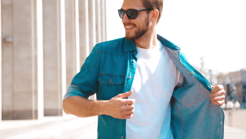 Portrait of handsome smiling stylish hipster lumbersexual model. Man dressed in jeans jacket clothes. Fashion male posing on the street background in sunglasses. Slow motion Royalty-Free Stock Footage #1029075503