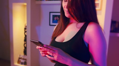 Close up of sexy Caucasian young woman with big boobs and black decorative dress using smartphone at home. Neon lights interior. 10Bit ProRes Color Graded. Camera pan rotating right to left.