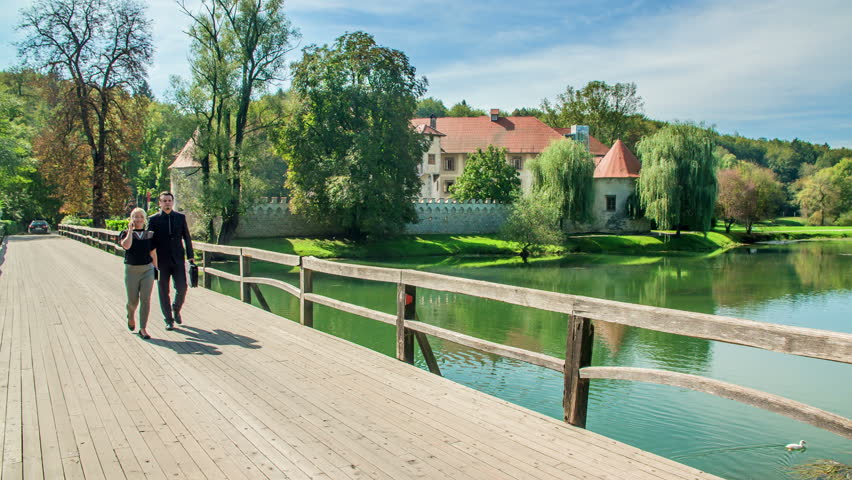 Two business people, a man and a woman are walking across a bridge. A young woman is talking on the phone and a man is carrying a black bag.