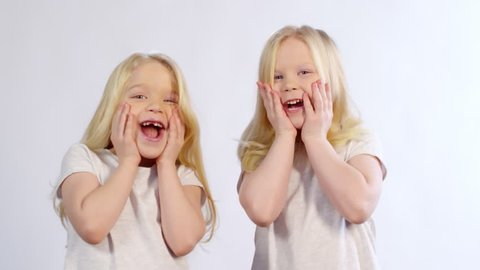 Waist-up shot of two 5-year-old twin girls with long blond hair, wearing plain t-shirts, slapping their hands to cheeks simultaneously and opening their mouths in surprise, on white background
