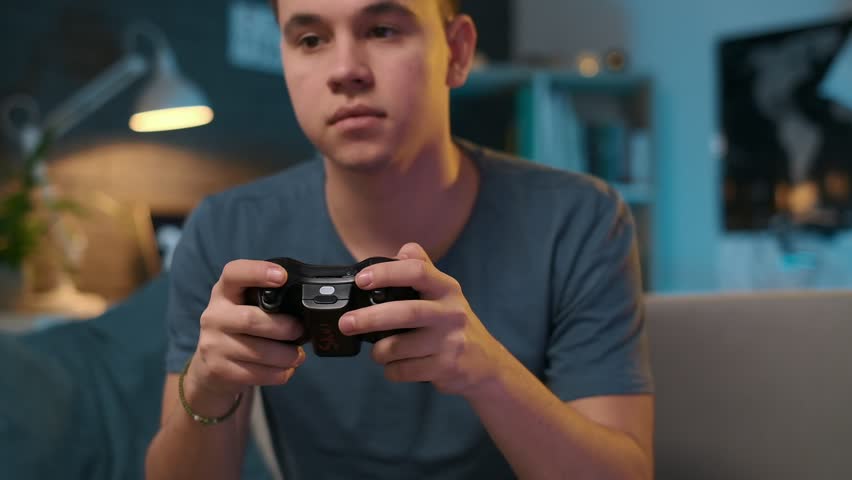 A hadsome teenager is sitting on a sofa and playing a video game on a playstation at home. Stay at home