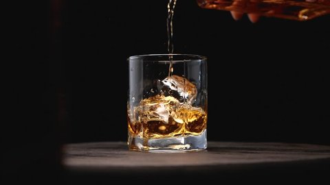 Glass of whiskey with ice cube. Alcohol pouring in the glass from the bottle. Scotch on the rocks. Slider shot. Dark, rustic, background.
