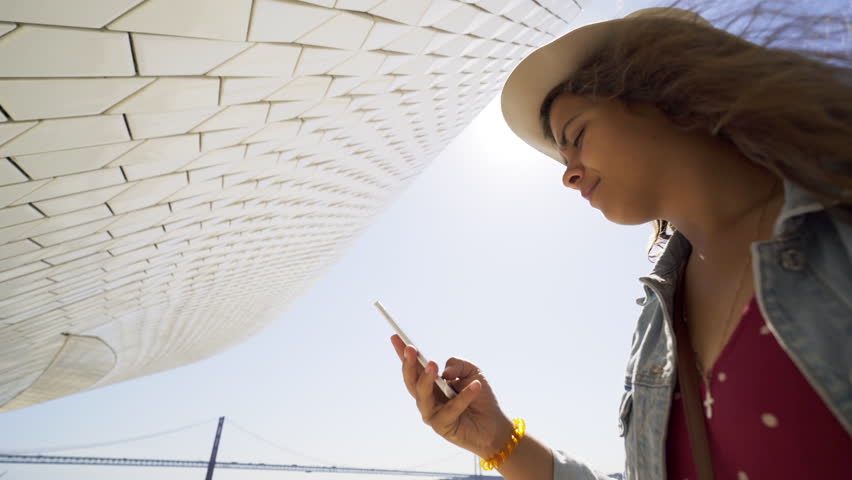 From below young lady taking selfie on mobile phone near modern buildings on embankment in Lisbon