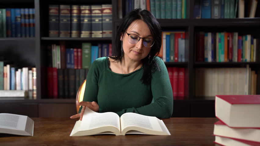 Beautiful young librarian woman sitting at wooden table and reading book in local library