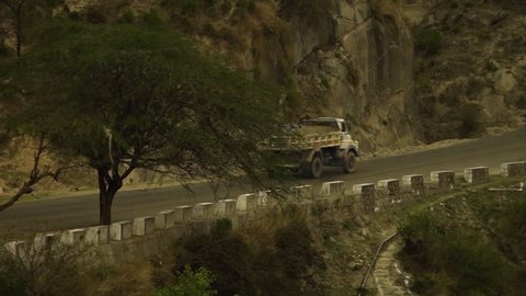 Seveal orange vehicles drive along a mountain road between a stone wall and a tall cliff face, while a cow runs from a man down the road