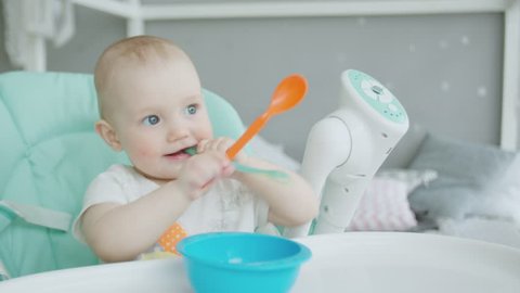 Close-up of teether toddler girl sitting on feeding chair in front of empty plate and gnawing plastic spoon. Cute infant teething and biting spoon before eating on chair for babies in children's room.
