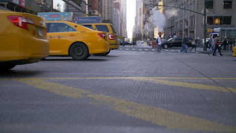 NEW YORK CITY, USA - 03/08/2019: New York city traffic commuting at rush hour. Slow motion Classic Yellow Taxi Cabs passing with Tourist and commuters walking on the sidewalk with smoke rising. 