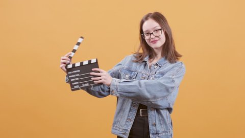 Beautiful teenage girl hold a clapperboard and smiles to the camera. Young girl in denim jacket over yellow background.
