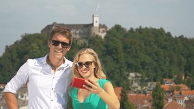 CLOSE UP, DOF: Happy Caucasian man kisses his girlfriend on the cheek while taking selfies on a rooftop in the sunny Ljubljana city center. Cheerful couple shooting a video for the traveling blog.