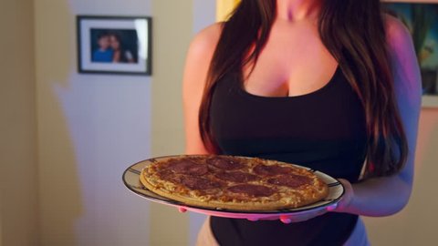 Close up video of a very pretty woman with sexy black t-shirt, holding a pizza in her hands at home. Neon lights interior. 10Bit ProRes Color Graded. Camera pan rotating to Right. 