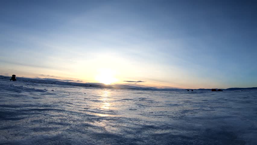 Winter sunrise blazes in the background as a fisherman drills a hole in a frozen lake to prepare to ice fish.  A popular sport in the colder states and countries.