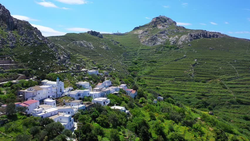Aerial drone video of picturesque traditional village of Koumaros in the slopes of mountain and castle of Exomvourgo or Exombourgo with beautiful deep blue sky, Tinos island, Cyclades, Greece