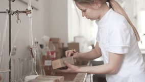 A close up video of a technologist who opens a carton box with the content. She uses a little spoon to take the earth out and pours it into the white plastic containers. The background is blurred