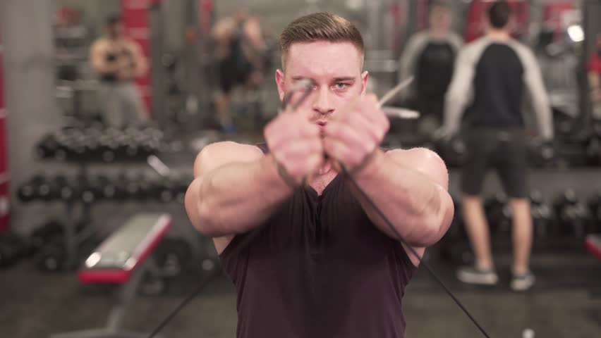 Portrait of Young Caucasian Male Bodybuilder in Gym Exercize Biceps on Cable Machine. Blurred Background WIth Other Gym Customers