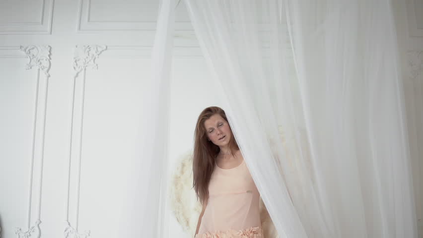 Young woman in cream dress wearing white wings, happy female model as angel smiling standing on bed with canopy in white hall studio and posing at camera