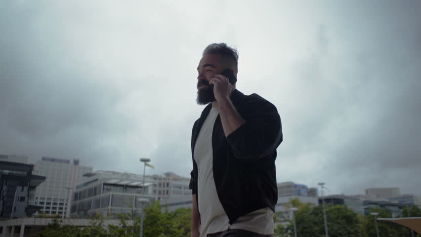 Relaxed man having pleasant conversation through smartphone. Handsome bearded brunette walking on city street and talking on phone during windy day. Communication and technology concept