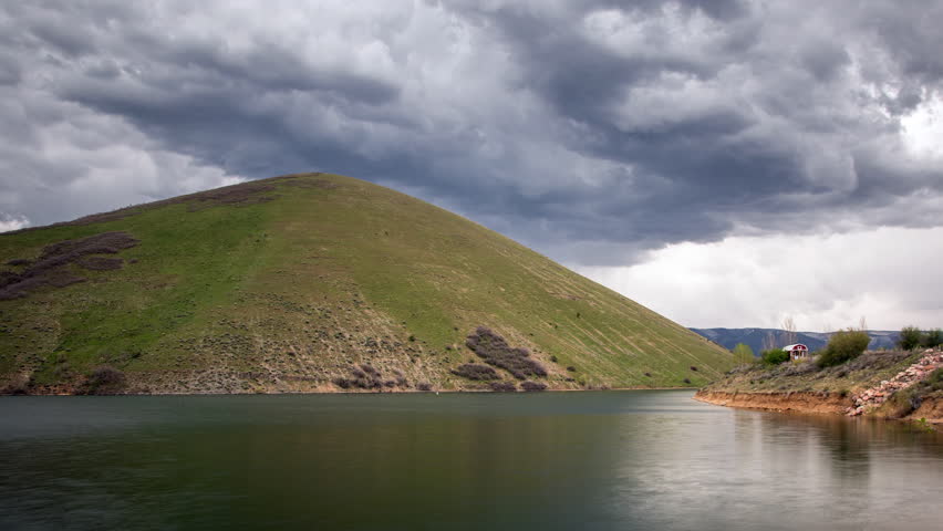 Time lapse of thick dark clouds moving Deer Creek Reservoir past grassy green hill in Utah.