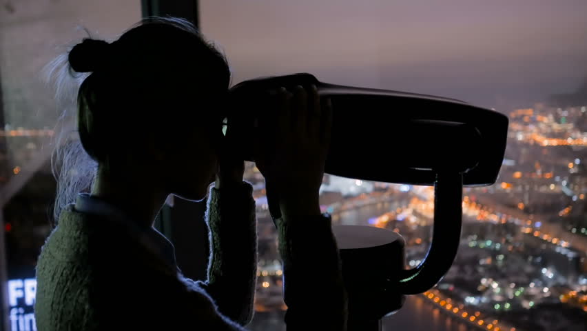 Young woman looking through tourist telescope from skyscraper, exploring night cityscape. Light city street bokeh illumination at evening. Relax, discover and journey concept