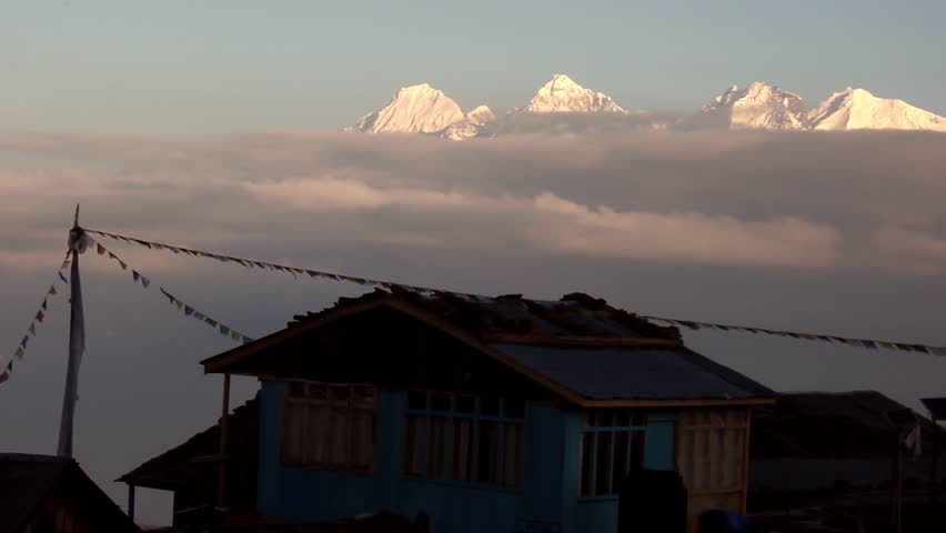 Epic view of hut with prayer flags sitting atop mountain above clouds with snow capped mountain peaks in the background in Nepal