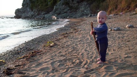 Cute 1,5 years old boy is playing with a big stick on sandy beach of seaside. Nakhodka, Russia. September evening
