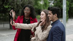 Side view of three young people standing in street, having video chat on tablet, Caucasian girl in camel trench presenting friends, they waving hand, greeting. Communication, lifestyle concept