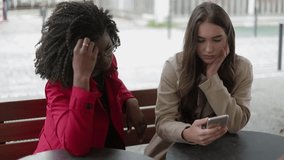 Pretty Afro-american woman in rose coat and attractive Caucasian girl in trench sitting outside, swiping photos on phone, discussing them. Wedding preparation, house searching. Communication concept