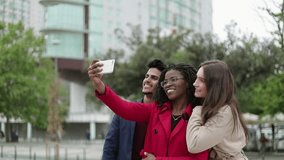 Pretty Caucasian and Afro-american women in camel and rose coat and serious mixed-race man in navy blue suite making selfie outside, posing, smiling. Lifestyle concept 