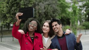 Pretty Caucasian and Afro-american women in camel and rose coat and serious mixed-race man in navy blue suite making selfie outside, hugging, posing, smiling. Lifestyle concept 