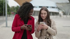 Pretty Afro-american woman in rose coat and attractive Caucasian girl in camel trench walking along street, one showing pictures on phone, they discussing, laughing. Communication, lifestyle concept