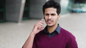 Front view of handsome young mixed-race man in dark rose pullover talking on phone outside, clarifying something, looking serious. Communication, work concept 