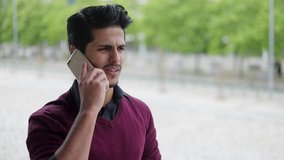 Medium view of handsome young mixed-race man in dark rose pullover talking on phone outside, clarifying something, looking serious. Communication, work concept 
