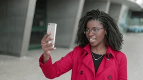 Medium view of pretty young Afro-american woman in spectacles and rose coat having video chat on phone outside, behaving emotionally, gesticulating. Communication, work concept 