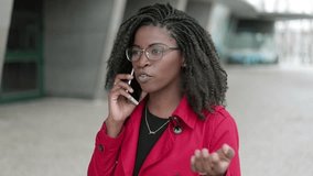 Medium view of pretty young Afro-american woman in spectacles and rose coat talking on phone outside, clarifying something, looking serious. Communication, work concept 
