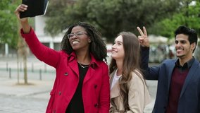 Pretty Caucasian and Afro-american women in camel and rose coat and smiling mixed-race man in blue suite standing one by one, making selfie on tablet outside, having fun, laughing. Lifestyle concept 