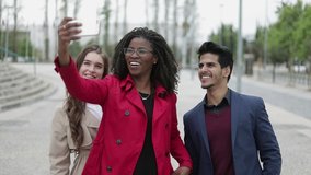 Pretty Caucasian and Afro-american women in camel and rose coat and smiling mixed-race man in navy blue suite making selfie outside, posing, smiling. Dolly shot. Lifestyle concept 
