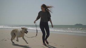 Smiling young Caucasian woman having fun with dog outdoor. Attractive young brunette with long hair running on sandy beach. Pet concept