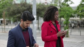 Side view of pretty Afro-american woman in spectacles and rose coat and handsome mixed-race man in navy blue suit walking along street, texting on phones. Communication, lifestyle concept