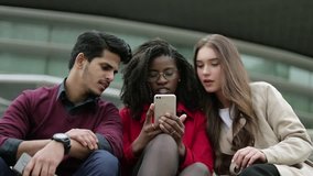 Front view of three young people sitting outside, googling on phone place to go, discussing, gesticulating. Leisure, lifestyle concept
