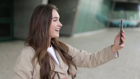 Medium view of attractive Caucasian girl in camel trench having video chat on phone outside, waving hand, showing surroundings, smiling. Side view. Communication, work concept 