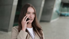Medium shot of attractive Caucasian girl in camel trench standing outside, talking on phone, flirting. Lifestyle, communication concept