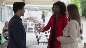 Side view of pretty Afro-american woman in rose coat shaking hands with serious mixed-race man, making deal, attractive Caucasian girl in trench standing near, they talking. Communication concept