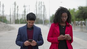 Pretty Afro-american woman in spectacles and rose coat and handsome mixed-race man in navy blue suit walking along street, texting on phones. Communication, lifestyle concept