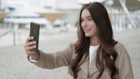 Medium view of attractive Caucasian girl in camel trench having video chat on phone outside, talking, gesticulating, sending kisses. Communication, work concept 